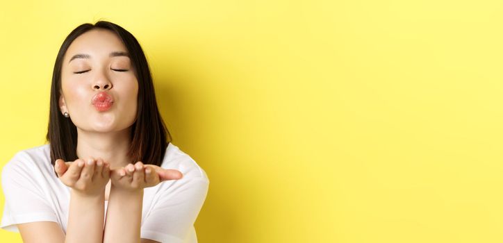 Valentines day concept. Close up of beautiful asian woman pucker lips and holding hands near mouth, blow air kiss at camera with closed eyes, standing over yellow background.