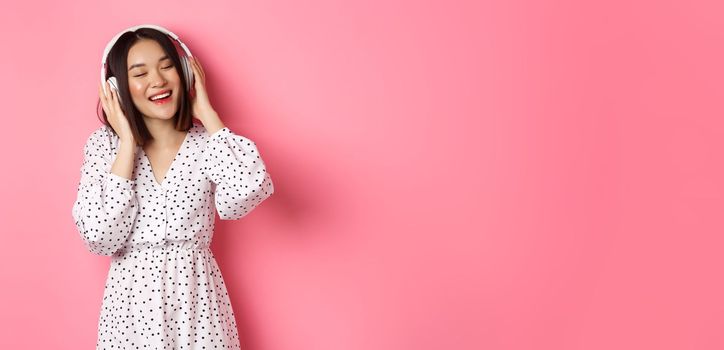 Romantic asian woman smiling happy, listening music in headphones and dancing, standing in trendy dress over pink background.