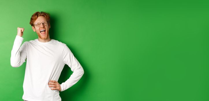 Young cheerful guy scream of joy, winning prize and celebrating, making fist pump gesture and smiling happy, standing over green background.