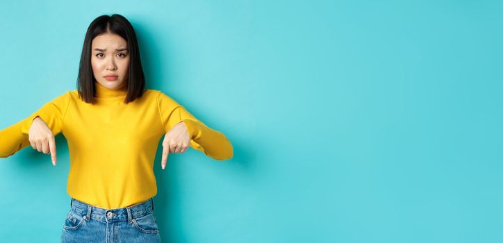 Shopping concept. Sad and worried asian girl frowning silly, pointing fingers down at mistake, standing upset over blue background.