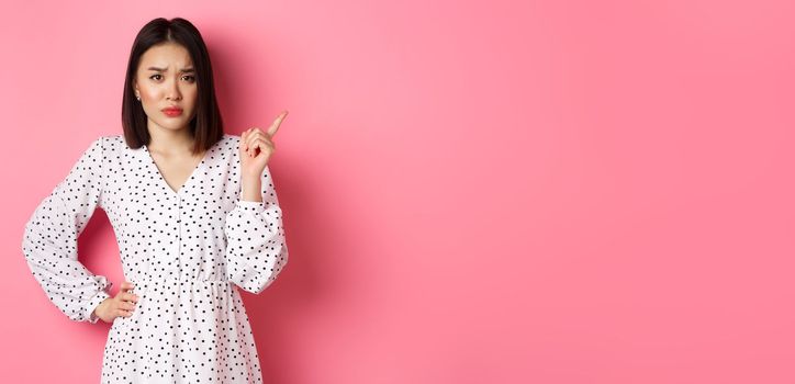 Sad and gloomy asian girl frowning, pointing finger at upper right corner and staring at camera unhappy, standing over pink background.