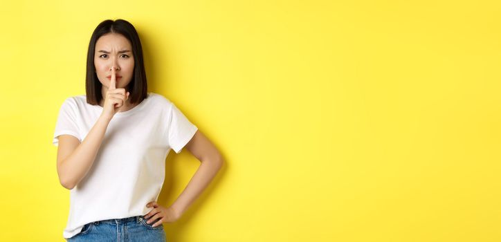 Beauty and fashion concept. Angry and bossy asian girl telling to be quiet, scolding loud people, shushing and frowning at camera, standing in t-shirt over yellow background.