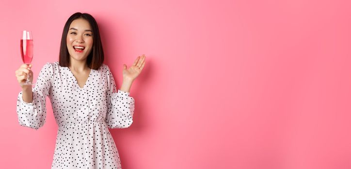 Happy asian woman celebrating, saying toast on party, raising glass of champagne and smiling, standing in dress over pink background.