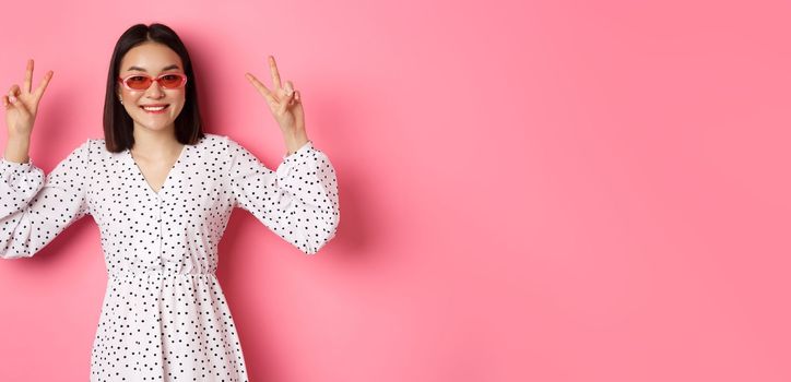 Tourism and lifestyle concept. Beautiful asian woman on summer vacation, showing peace signs and smiling happy, wearing trendy sunglasses, pink background.