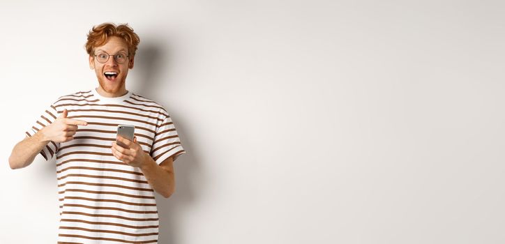 Technology and e-commerce concept. Amazed redhead man checking out online promo offer on mobile phone, pointing finger at smartphone and smiling, white background.