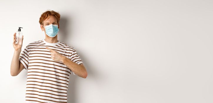Covid-19, health and lifestyle concept. Cheerful redhead man in face mask pointing finger at hand sanitizer, recommending antiseptic, white background.