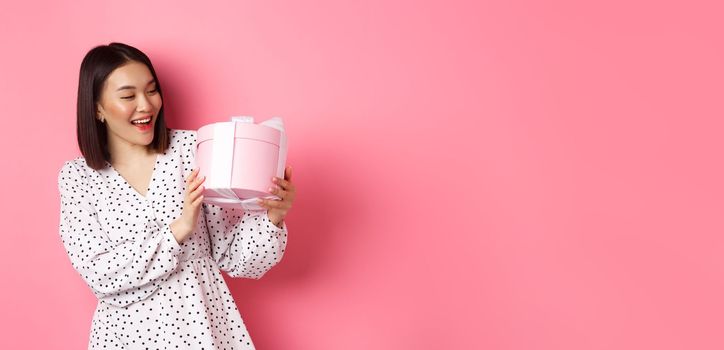 Valentines day, celebration concept. Beautiful asian woman holding romantic gift box, smiling happy, standing over pink background.