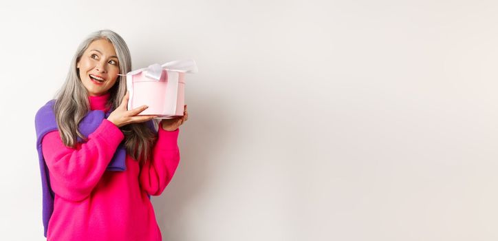 Holidays and valentines day concept. Happy asian mature woman shaking box with gift, guessing what inside present, standing over white background.