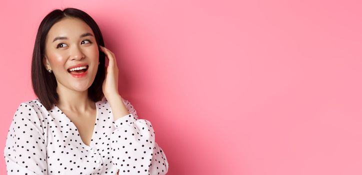 Close-up of flirty asian woman sighing, looking dreamy left and smiling, standing over pink background. Copy space