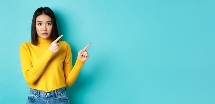 Shopping concept. Disappointed korean girl looking gloomy, asking to buy this, pointing fingers at upper right corner and staring sad at camera, blue background.