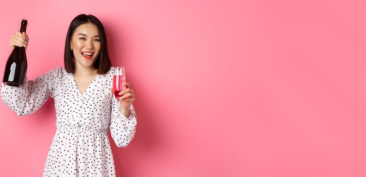 Happy asian woman celebrating, having fun and partying, pouring glass of champagne and laughing, standing over pink background.