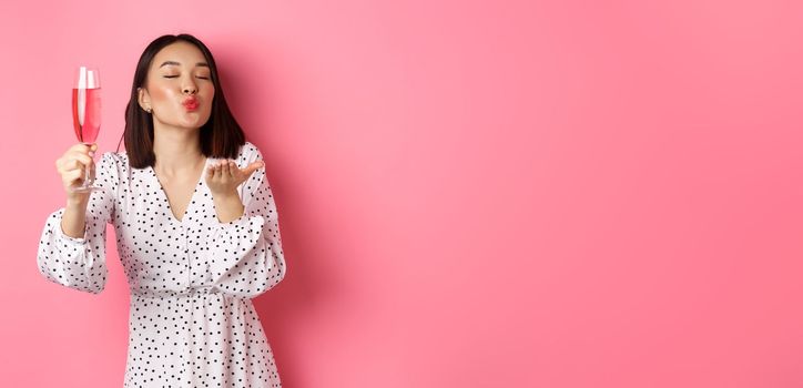 Romantic asian woman raising glass of champagne and sending air kiss at camera, celebrating and having fun, standing over pink background.