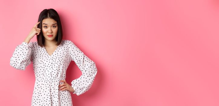 Image of attractive asian lady scolding person, pointing at head and staring at camera disappointed, are you stupid gesture, standing over pink background.