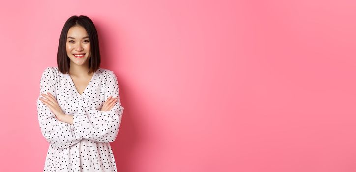 Pretty young asian woman in spring dress smiling, cross arms on chest and looking confident, standing over pink background.