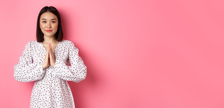 Beautiful asian lady in dress asking for help, holding hands in pray or namaste gesture, thanking you, standing over pink background.