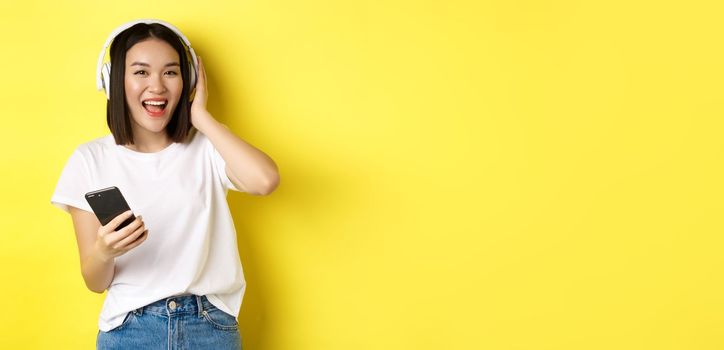 Cool asian girl dancing and listening music in wireless headphones, holding smartphone in hand, standing over yellow background.