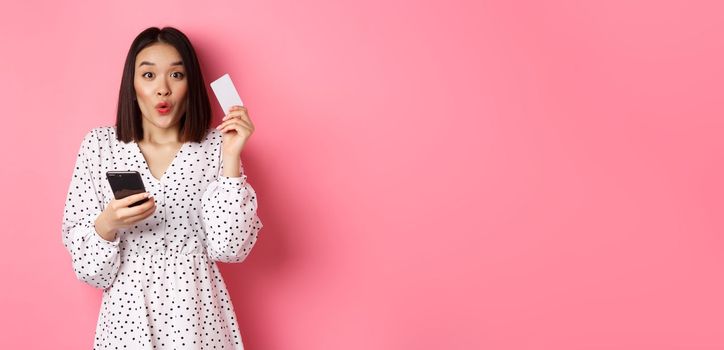 Attractive young asian woman order online, holding credit card and mobile phone, making internet purchase, standing happy over pink background.