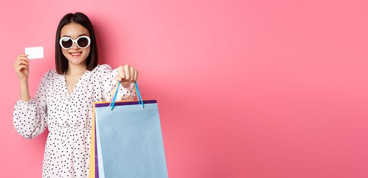 Beautiful asian woman in sunglasses going shopping, holding bags and showing credit card, standing over pink background.