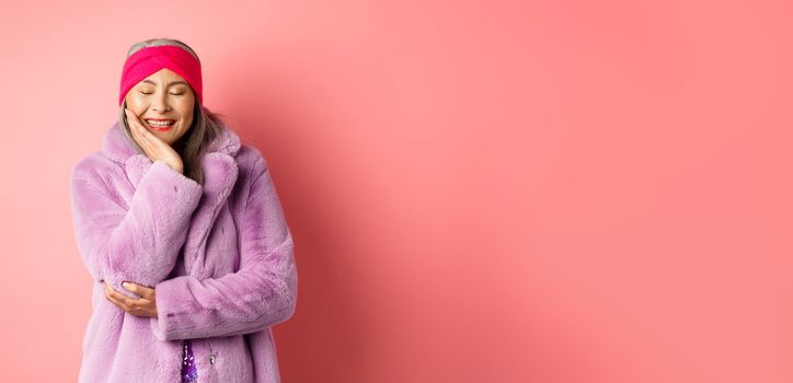 Fashion and shopping. Happy asian mature woman in purple faux fur coat smiling lovely and carefree, close eyes with happiness, standing against pink background.