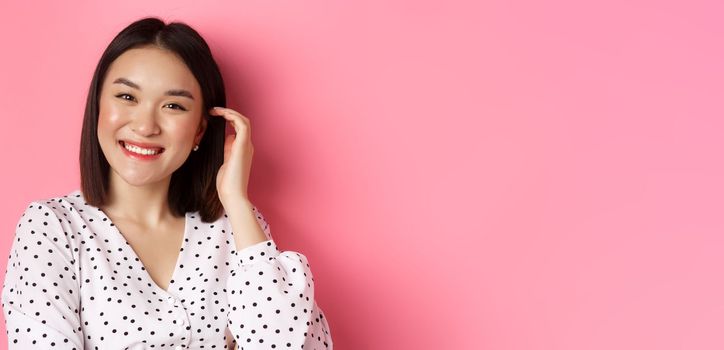 Close-up of beautiful asian woman smiling happy, touching new haircut, standing over pink background.