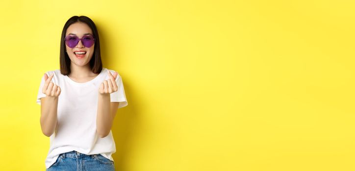 Fashion and lifestyle concept. Attractive asian woman in heart-shape sunglasses, showing finger hearts and smiling happy at camera, standing over yellow background.