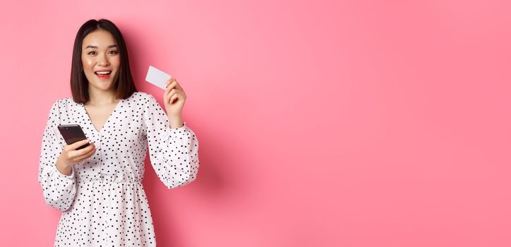 Attractive young asian woman order online, holding credit card and mobile phone, making internet purchase, standing happy over pink background.