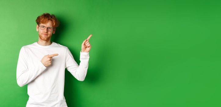 Skeptical caucasian man in glasses having doubts, pouting and frowning while pointing fingers right at something bad, standing over green background.