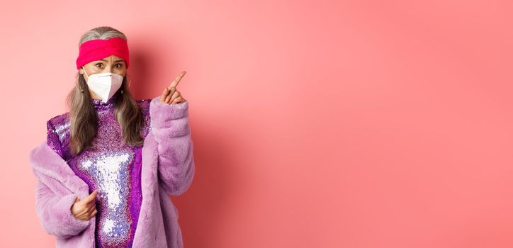 Coronavirus and shopping concept. Worried asian senior woman in stylish outfit and respirator pointing upper right corner, looking unsure at camera, asking advice, pink background.