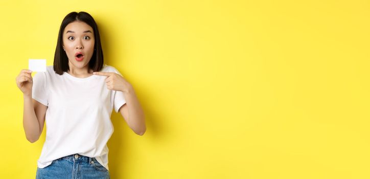 Young asian woman in casual white t-shirt showing plastic credit card and smiling at camera, yellow background.