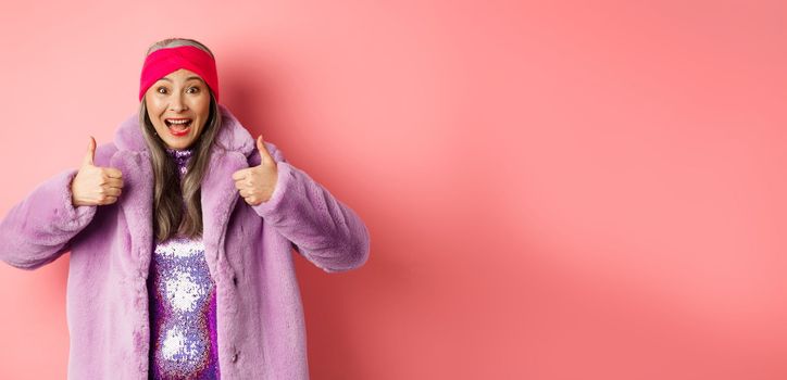 Fashion and shopping concept. Cheerful asian old woman showing thumbs-up in approval, like and recommend promotion, standing in stylish purple fur coat, pink background.