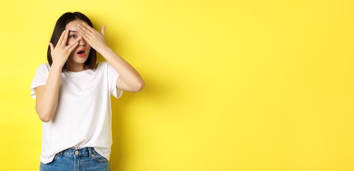 Intrigued asian girl waiting for surprise, peeking through fingers on eyes, smiling happy, standing over yellow background.