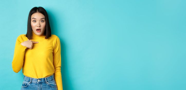 Image of surprised asian girl gasping and pointing at herself with disbelief, standing over blue background.