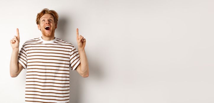 Fascinated redhead man looking with amazement and happiness, pointing fingers up, checking out something cool, standing over white background.