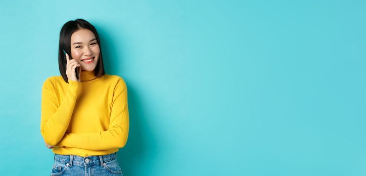 Young stylish asian woman talking on phone, calling friend and smiling, standing with smartphone against blue background.