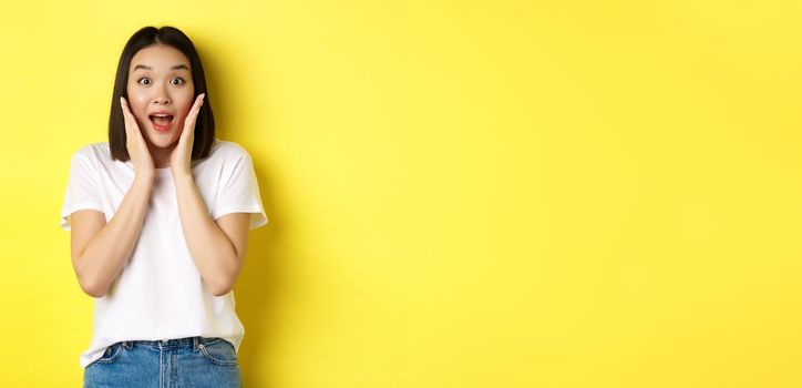 Beauty and fashion concept. Surprised asian girl looking amazed at camera, saying wow, checking out advertisement, standing over yellow background.