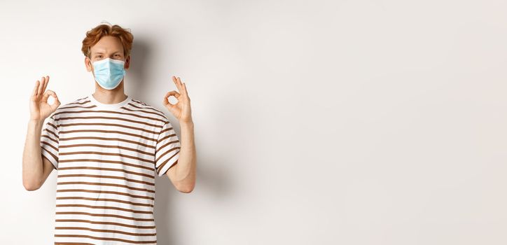 Covid-19, pandemic and social distancing concept. Young man with red hair wearing medical mask to prevent catching coronavirus, showing okay signs in approval, white background.