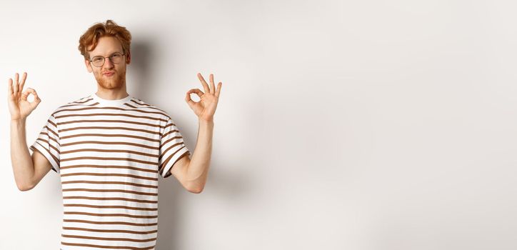 Attractive redhead guy in glasses showing okay signs and looking impressed, praising something cool, standing over white background.