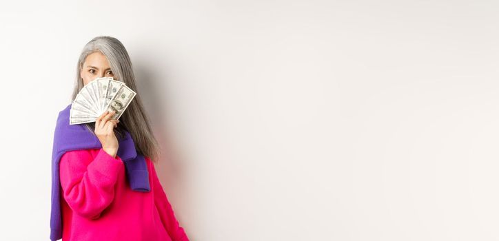 Shopping concept. Rich and fashionable korean senior woman hiding face behind money, looking at camera, standing over white background.