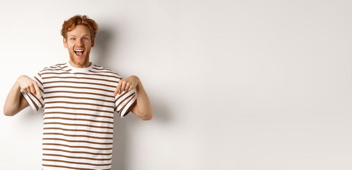 Enthusiastic young man with ginger hair and bristle showing advertisement, pointing fingers down and smiling, white background.