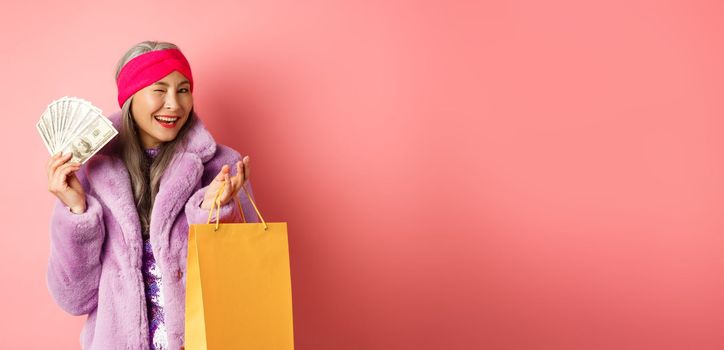 Rich and fashionable asian senior woman wasting money in shops, holding shopping bag and dollars, winking happy at camera, pink background.