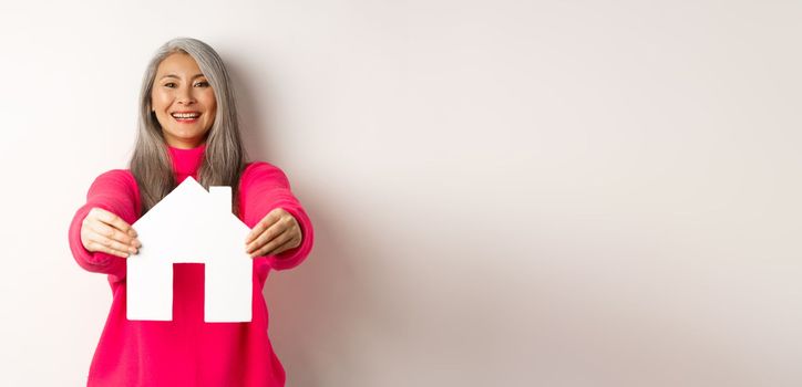 Real estate. Happy korean middle-aged woman with grey hair, showing paper house model and smiling, standing in pink sweater over white background.