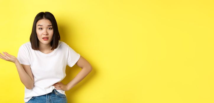 Careless asian woman asking so what, raise hand up and staring skeptical at camera, standing over yellow background.