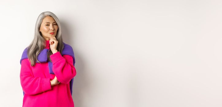 Image of thoughtful asian senior woman squinting suspicious at camera, having assumption, standing over white background and thinking.