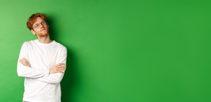 Confident young man with red hair, wearing glasses, looking smug at right side copy space, cross arms on chest self-assured, standing over green background.