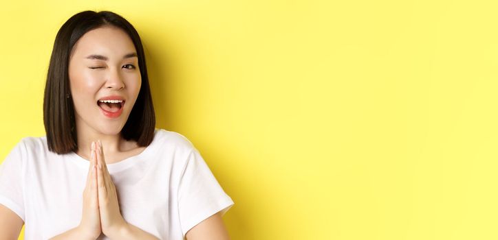 Close of cheeky young asian woman holding hands in namaste, thank you gesture, winking at camera coquettish, feeling lucky, standing over yellow background.