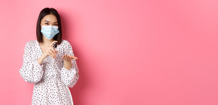 Covid-19, pandemic and lifestyle concept. Cute asian woman clean hands with sanitizer, using antiseptic and wearing medical mask, standing over pink background.