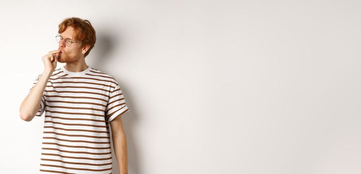 Image of satisfied young man with red hair and glasses kissing fingers, showing chefs kiss gesture to praise something perfect, standing over white background.