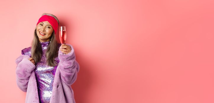 Gorgeous asian senior female in stylish purple faux fur coat and glittering dress, raising glass of champagne in cheers gesture, saying toast on party, pink background.