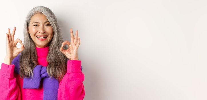 Close-up of satisfied asian female with grey hair, smiling pleased and showing OK signs, approve and like product, praising something awesome, white background.