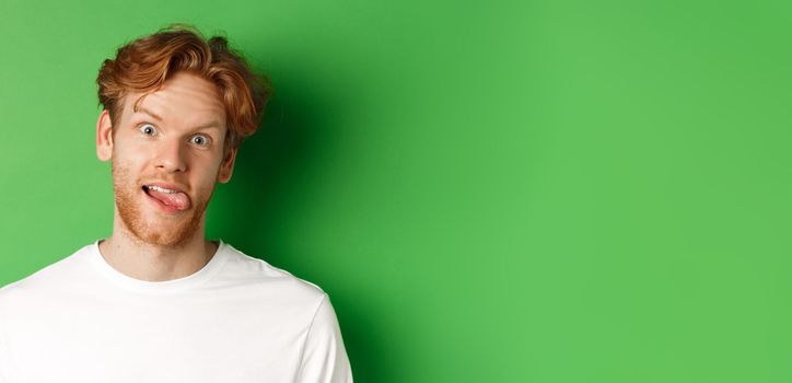 Emotions and fashion concept. Close up of funny redhead man showing silly faces, sticking tongue and staring at camera, standing over green background.